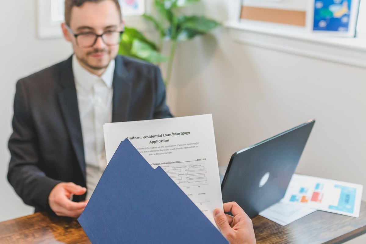 Hands presenting a residential loan application folder across a desk to a banker in a suit